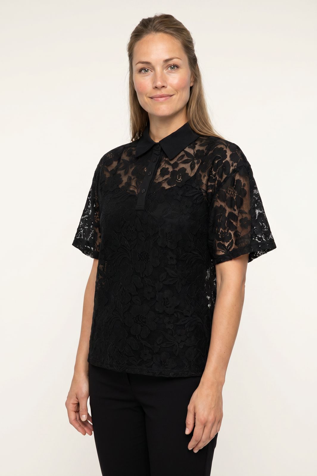 Kvinde iført en sort kortærmet blonde-poloshirt med blomstermønster mod en hvid baggrund.